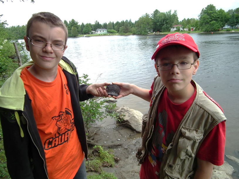 Two boys holding the same shard of pottery, as large as a hand. There is a body of water in the background.