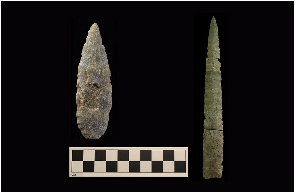 Two lance-shaped chipped stone points, with the one of the left wide and grey, and the one on the right ochre, long, and narrow. There is a photographic scale with black and white squares under the point on the left.
