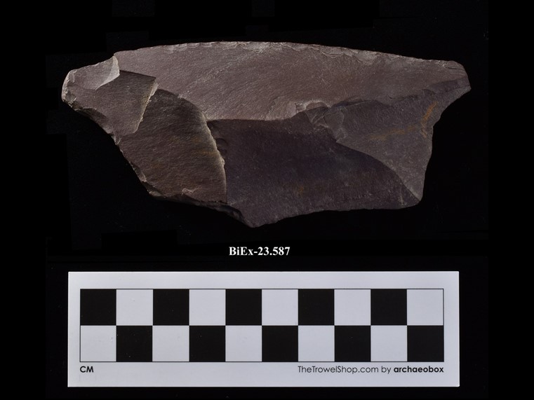 Chipped stone shard of a reddish colour, in an irregular polygon shape, of greater width than height. The top edge has been tapered. The number BiEx-23.587 is inscribed on the bottom. Below the image is a photographic scale in centimetres with black and white squares.