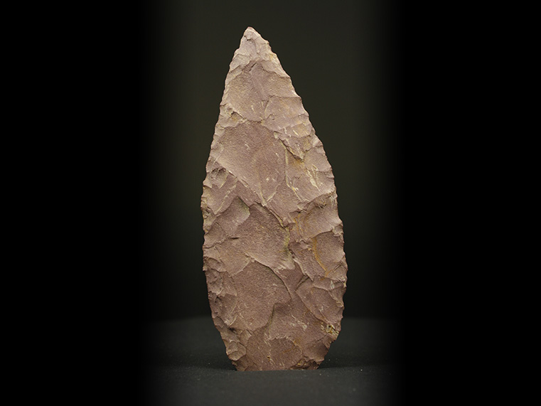 Two sides of a long, ochre chipped stone, divided into four fragments. It is lance-shaped with a convex base. The number BiEx-23.1839-1840-1841-1842 is inscribed underneath. Below the image is a photographic scale in centimetres with black and white squares.
