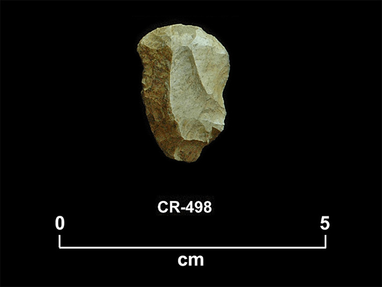 Fragment de pierre taillée beige de forme triangulaire inversée, aux coins arrondis et au front convexe. La cote CR-498 et une échelle 0 à 5 centimètres sont inscrites en dessous.