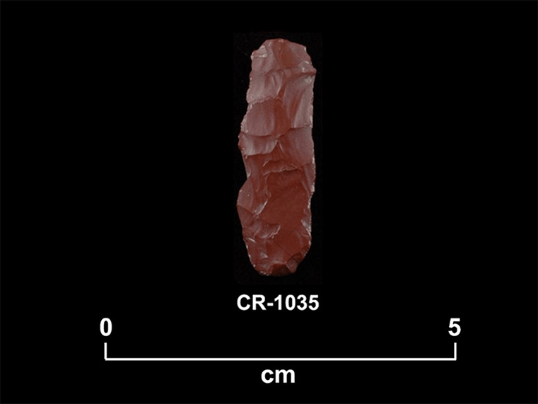 Pierre taillée rougeâtre de forme rectangulaire, orientée vers le haut. La cote CR-1035 et une échelle 0 à 5 centimètres sont inscrites en dessous.