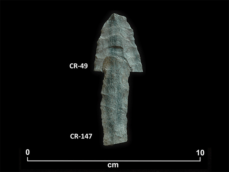 Two fragments of greenish chipped stone placed one above the other to form an arrow pointing up. The top fragment bears number CR-49, and the bottom fragment is CR-147. At the bottom, there is a scale of 0 to 10 centimetres.