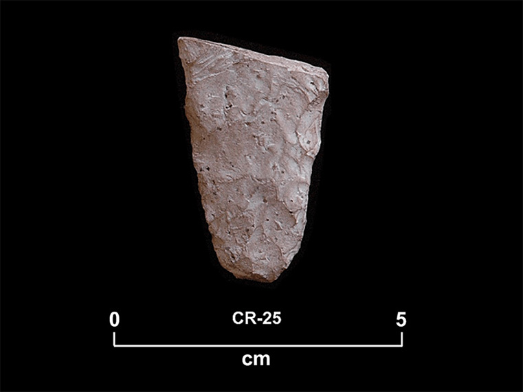 Fragment de pierre taillée altérée blanche à la base arrondie et aux bords droits divergents. La cote CR-25 et une échelle 0 à 5 centimètres sont inscrites en dessous.
