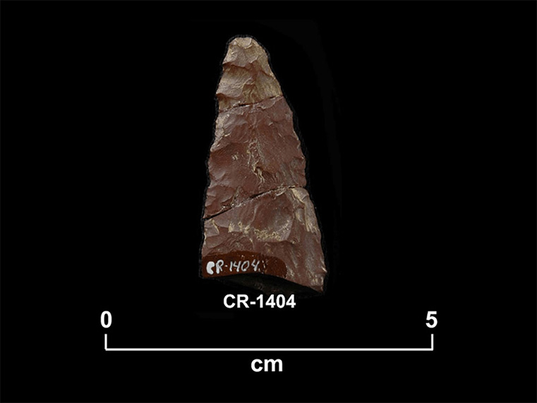Fragment de pierre taillée rougeâtre de forme triangulaire allongée, constitué de trois fragments recollés. La cote  CR-1404 et une échelle 0 à 5 centimètres sont inscrites en dessous.