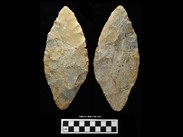 Two sides of a beige chipped stone composed of three fragments. The stone is lance-shaped. The number BiEx-23.1078-1230-1231 is inscribed on the bottom. Below the image is a photographic scale with black and white squares.
