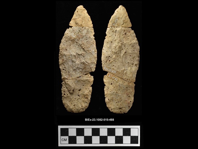 Two sides of a beige chipped stone composed of three fragments. The stone is oval and lance-shaped. The number BiEx-23.1082-515-488 is inscribed on the bottom. Below the image is a photographic scale with black and white squares.
