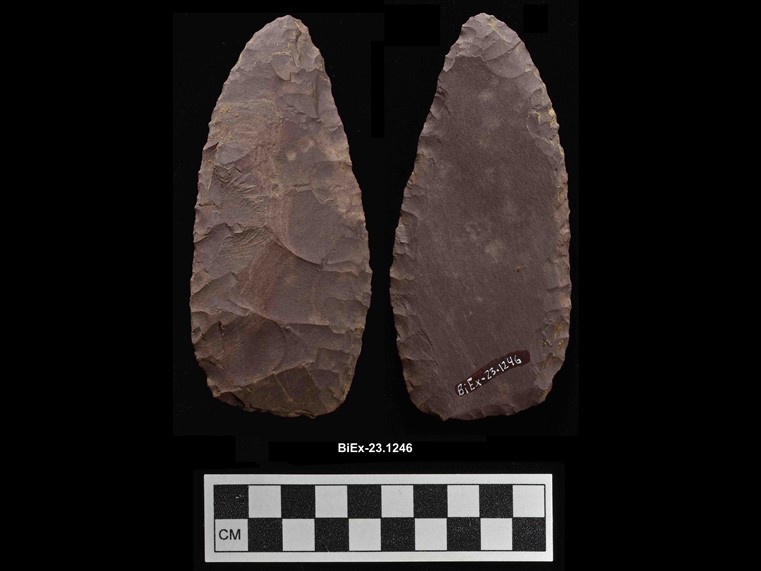 Two sides of a reddish, oval, lance-shaped chipped stone. The number BiEx-23.1246 is inscribed on the bottom. Below the image is a photographic scale with black and white squares.
