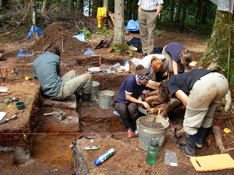 Deux carrés de fouille excavés avec, à l’intérieur, du matériel de fouille : seau, affichettes. En arrière-plan, des fouilleurs dans d’autres carrés de fouille d’un sous-bois. Il y a six personnes sur la photo.