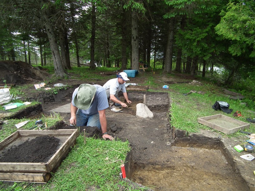 Two people crouching in an excavation square located in a wooded area. There are two sifting screens, one over the other, in the foreground and large trees with a picnic table in the background.
