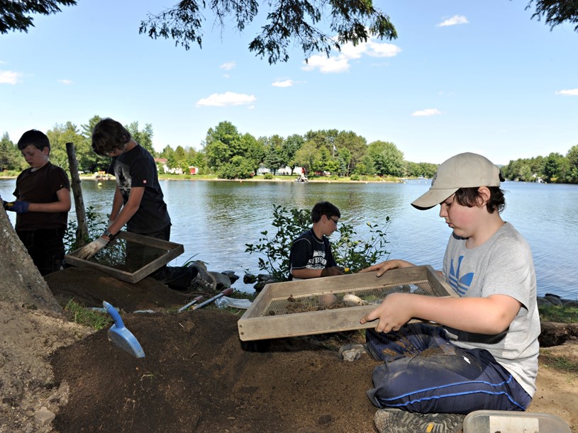 Four boys squatted on the ground. One of them in the foreground is holding a sifting screen. There is a body of water in the background.