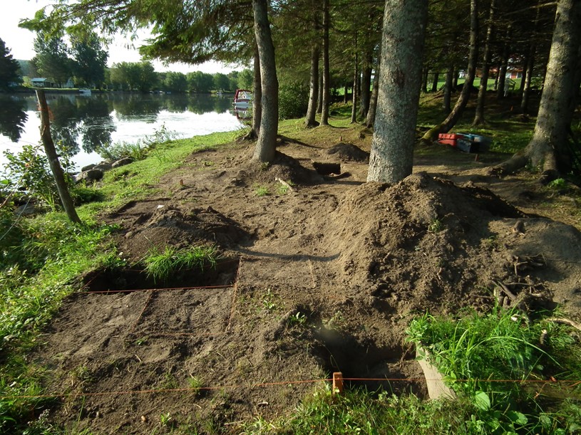 A weeded space in undergrowth at the edge of a body of water, with two square holes in the foreground, delineated by stakes and red string.