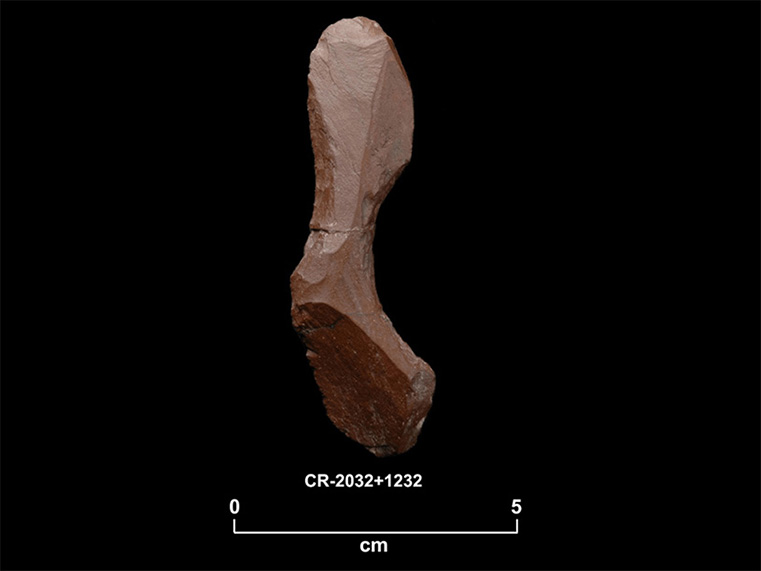 Fragment de pierre taillée rougeâtre en forme de L avec deux bords concaves. La cote CR-2032 + 1232 et une échelle 0 à 5 centimètres sont inscrites en dessous.