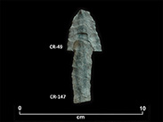 Two fragments of greenish chipped stone placed one above the other to form an arrow pointing up. The top fragment bears number CR-49, and the bottom fragment is CR-147. At the bottom, there is a scale of 0 to 10 centimetres.