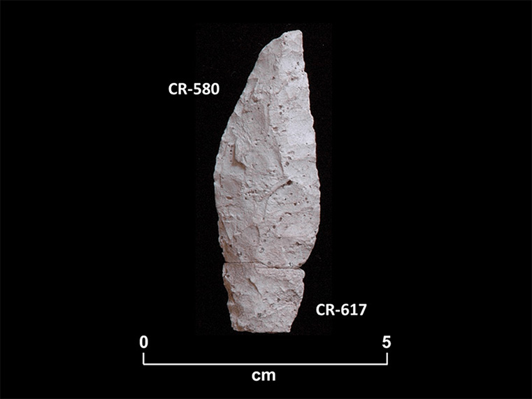 Deux fragments altérés d’une pointe en pierre taillée blanche. La base a une forme de pédoncule. La cote CR-580 est associée à la partie supérieure et la cote CR-617, à la partie inférieure. Une échelle 0 à 5 centimètres est inscrite en dessous.