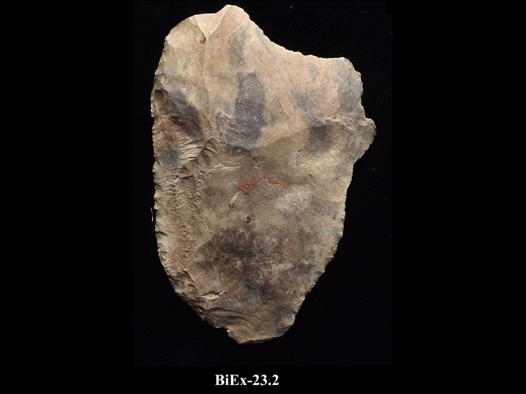 Beige chipped stone shard with an irregular polygon shape and a sharp edge. The number BiEx-23.2 is inscribed on the bottom.