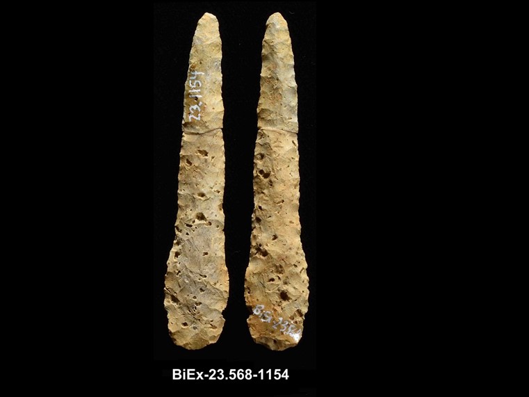 Two sides of a long, ochre-coloured chipped stone, divided into four fragments. The shape is linear. The number BiEx-23.568-1154 is inscribed on the bottom.