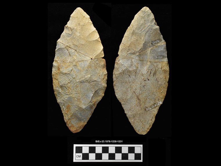 Two sides of a beige chipped stone composed of three fragments. The stone is lance-shaped. The number BiEx-23.1078-1230-1231 is inscribed on the bottom. Below the image is a photographic scale with black and white squares.