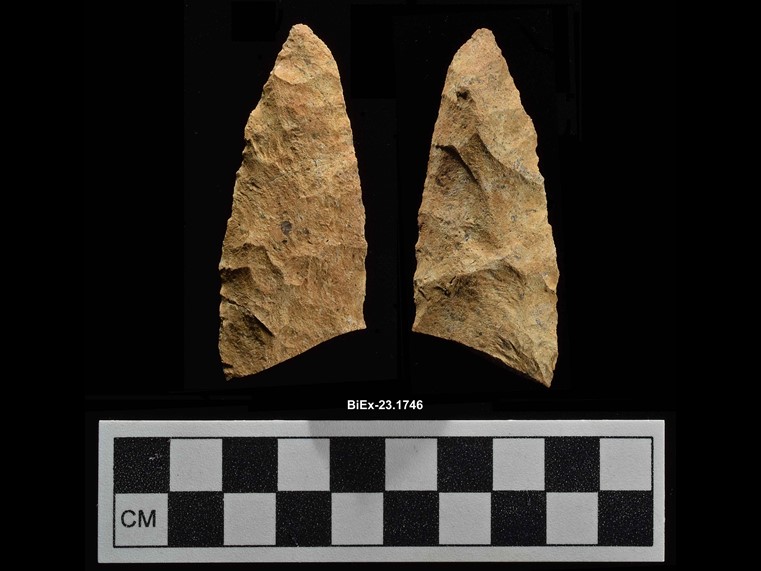 Two sides of a fragment of a beige chipped stone. The stone is lance-shaped, and its base is broken at an oblique angle. The number BiEx-23.1746 is inscribed on the bottom. Below the image is a photographic scale with black and white squares.