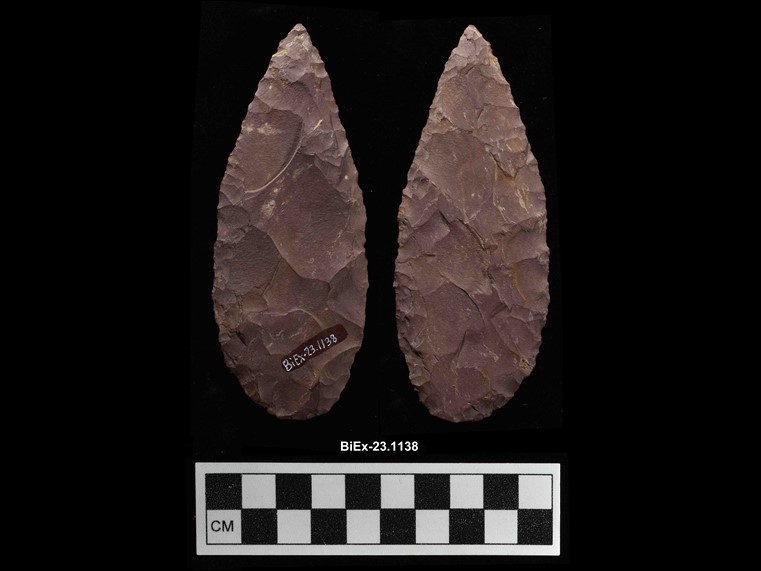 Two sides of a reddish, oval, lance-shaped chipped stone. There are visible marks of parts that were chipped off the edge of the stone. The number BiEx-23.1138 is inscribed in white on the bottom. Below the image is a photographic scale with black and white squares.