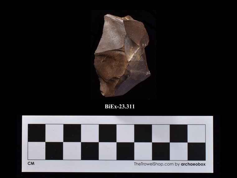 A block of reddish stone, more or less pyramidal, with a missing piece at its peak. The number BiEx-23.311 is inscribed on the bottom. Below the image is a photographic scale with black and white squares.
